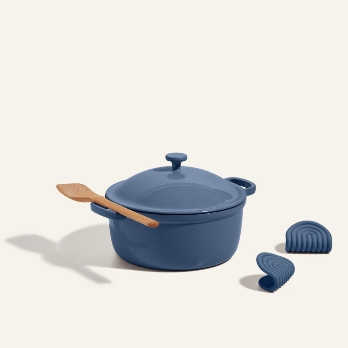 The Cast Iron Perfect Pot in blue, with its lid, a wooden spatula on top, and two matching blue silicone handle covers nearby, is displayed on a light background.