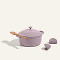 The lavender Cast Iron Perfect Pot with lid, a wooden spatula on top, and two silicone pot holders beside it is displayed against a neutral background.