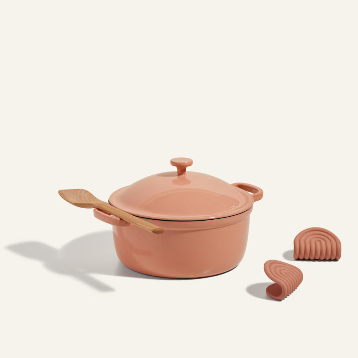 The Cast Iron Perfect Pot in pink with a ceramic finish comes with a lid, a wooden spoon resting on top, and two matching pink silicone pinch mitts beside it.