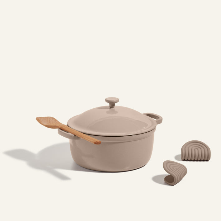 The Cast Iron Perfect Pot in beige, topped with a matching lid and wooden spatula, is displayed with two beige silicone mitts against a light background.