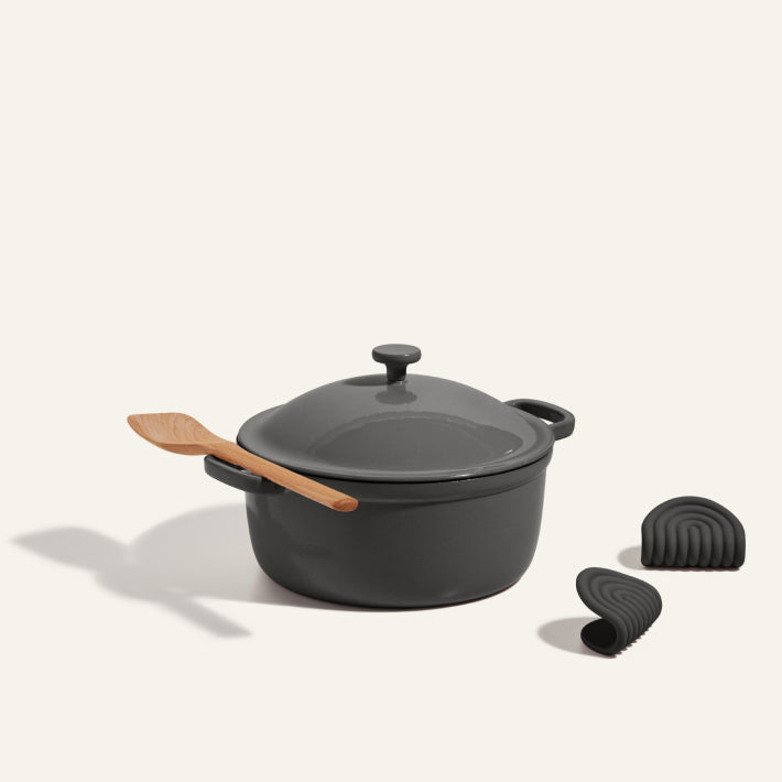 The Cast Iron Perfect Pot in dark gray with lid features a wooden spoon on top and comes with two black silicone pot holders, all displayed on a light background.