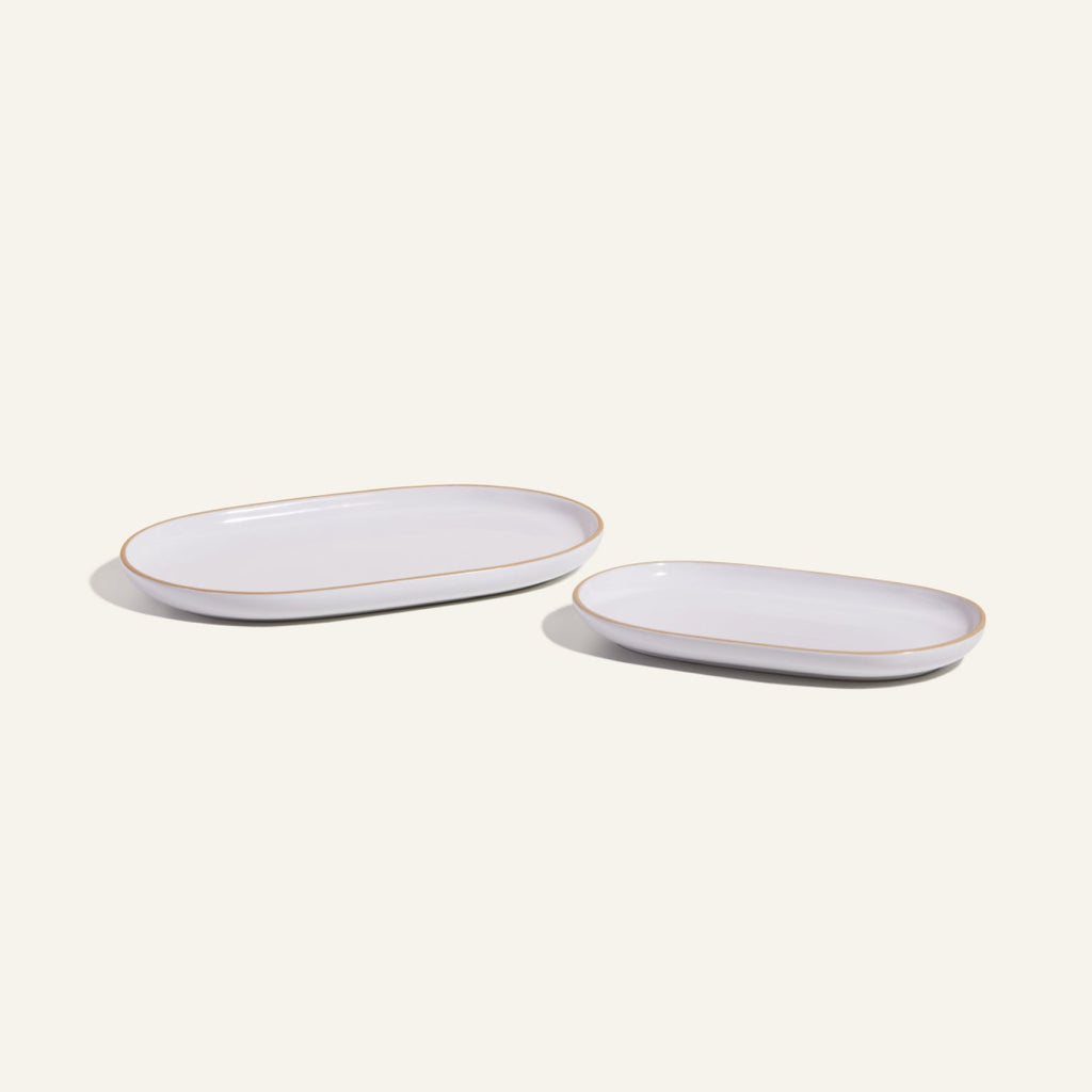 Two Serving Platters, one large and one small, feature an elegant white oval ceramic design with tan rims. Placed side by side on a light background, they showcase stylish and stackable tableware.