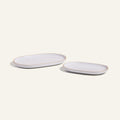 Two Serving Platters, one large and one small, feature an elegant white oval ceramic design with tan rims. Placed side by side on a light background, they showcase stylish and stackable tableware.
