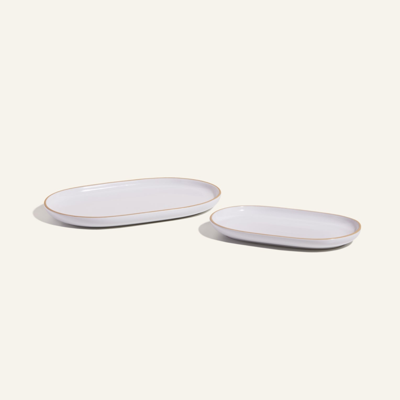 Two Serving Platters, one large and one small, feature an elegant white oval ceramic design with tan rims. Placed side by side on a light background, they showcase stylish and stackable tableware.