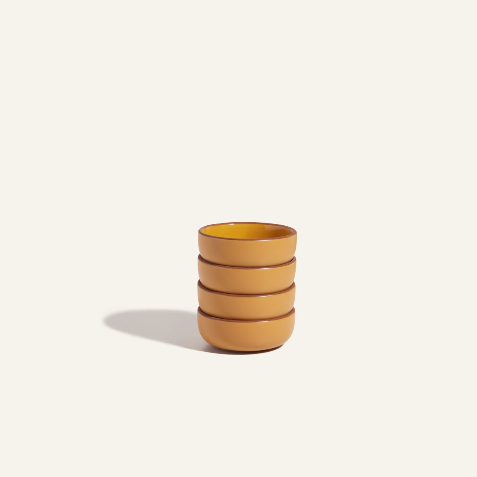 Four round, orange Tiny Bowls—stackable ceramic tableware—are arranged in a stack on a plain, light-colored surface with a shadow cast to the left.