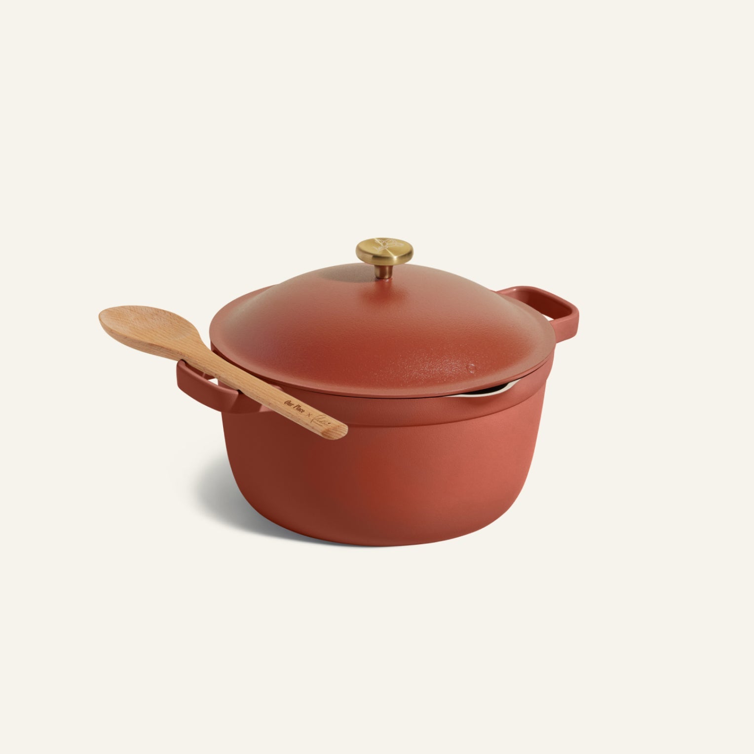The Perfect Pot, a red cast iron Dutch oven with lid and brass knob, features a wooden spoon resting on its handle, displayed against a plain light background.