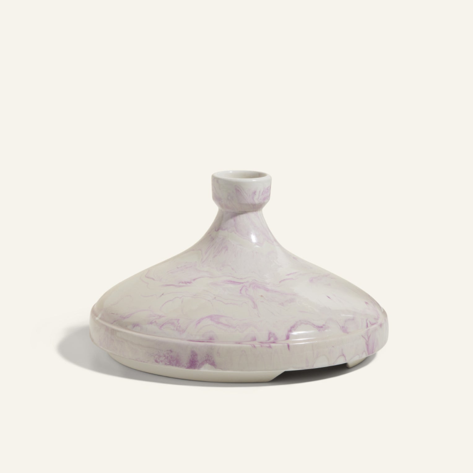 The Tagine is a handcrafted clay lid from Morocco featuring a marbled white and light purple pattern, ideal for slow cooking, shown against a plain light background.
