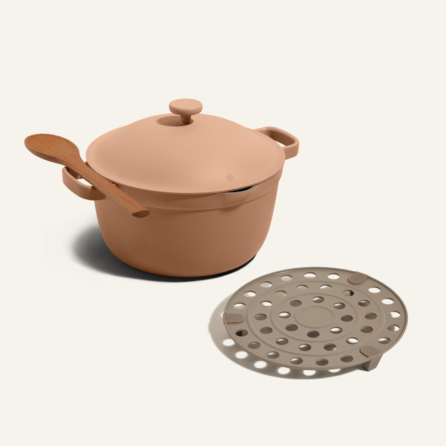 A Perfect Pot in beige with lid, a wooden spoon resting on its handle, and the Perfect Pot Roasting Rack positioned beside it.