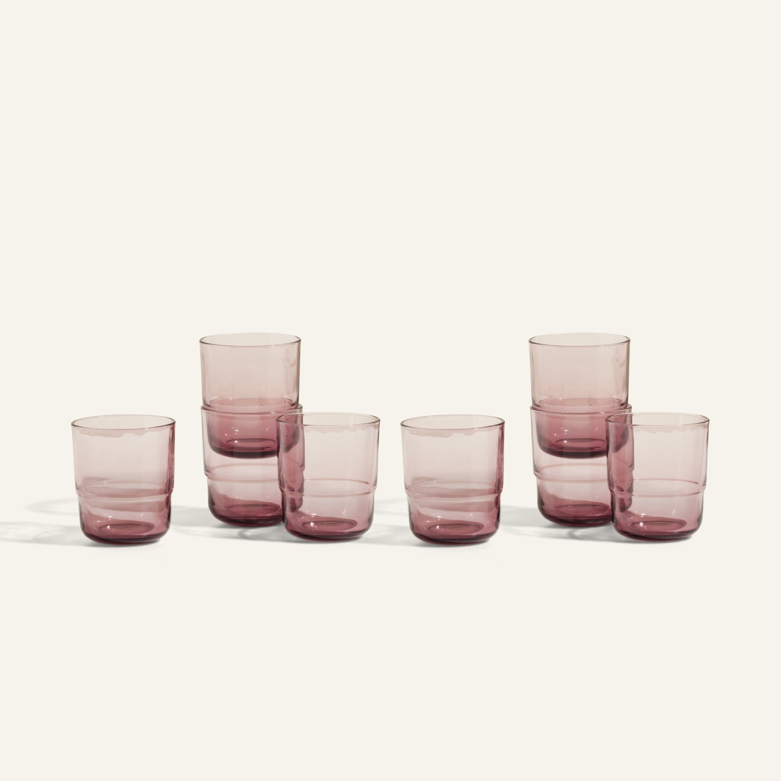 Seven handmade, transparent light purple Short Night + Day Glasses are arranged in a row on an off-white surface. Some are stacked in pairs and others stand alone, highlighting the versatility of this unique tumbler set.