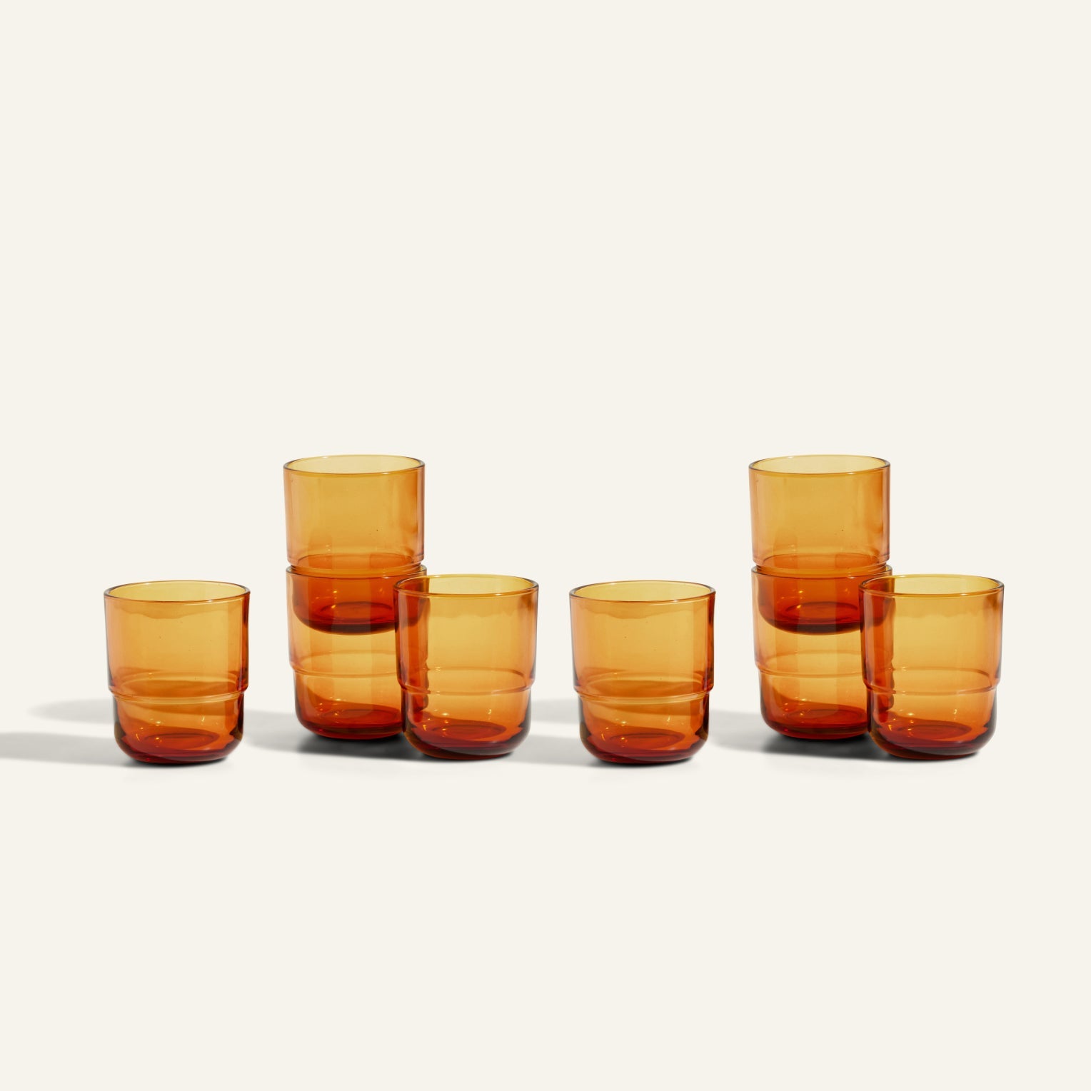 Six Short Night + Day Glasses made from amber recycled glass are displayed on a light surface—two stacked centrally, the rest standing alone. These versatile tumblers are perfect for any table setting.