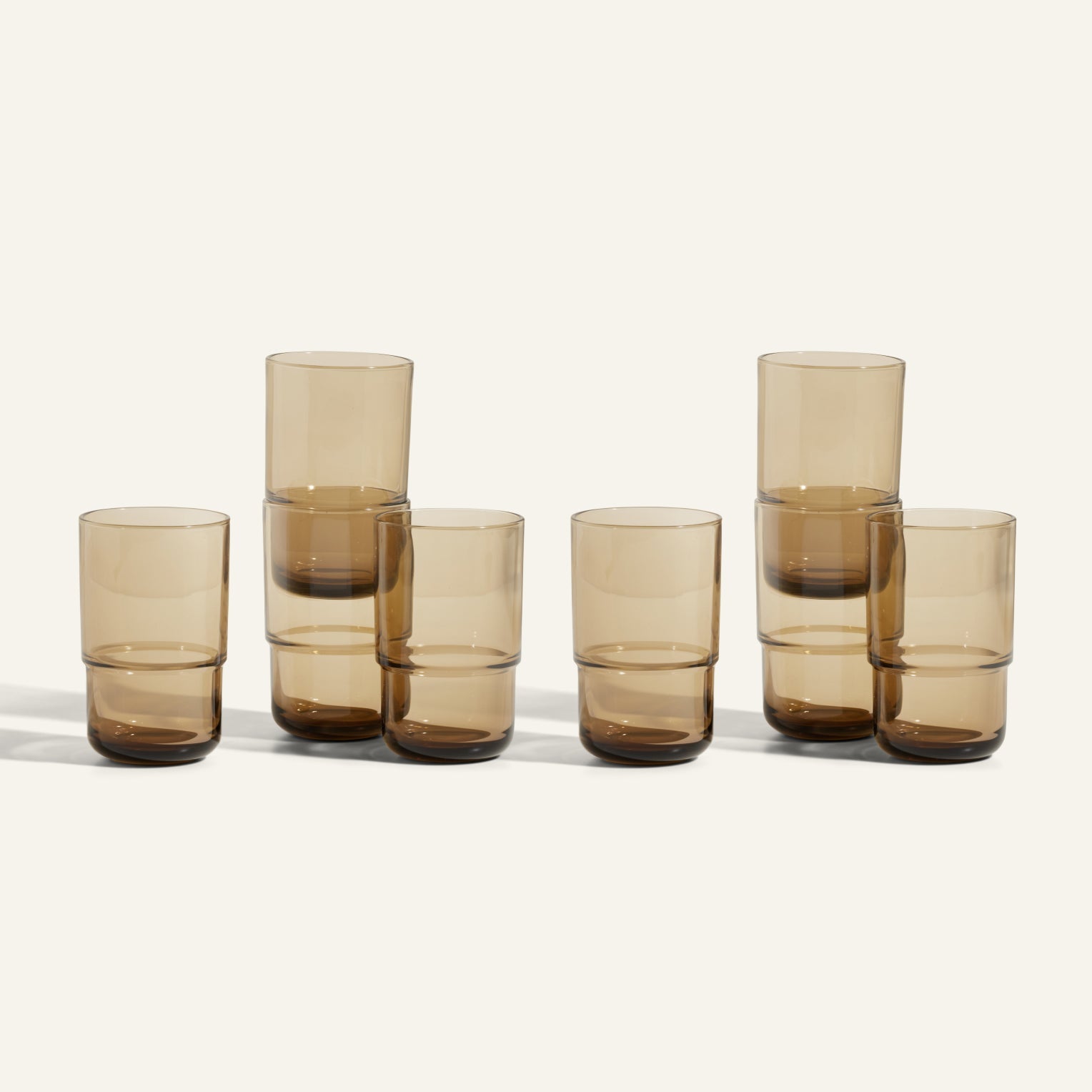 Seven Tall Night + Day Glasses, amber-colored and handmade with a 16 oz capacity each, are arranged in three uneven stacks on a light surface against a plain background.