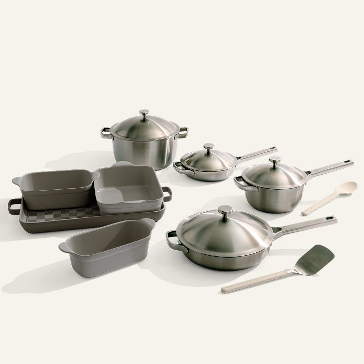 The Titanium Pro Cookware + Bakeware Set includes stainless steel pots with lids, pans, nonstick baking dishes and loaf pan, a wooden spoon, and metal spatula, all neatly displayed on a white surface.