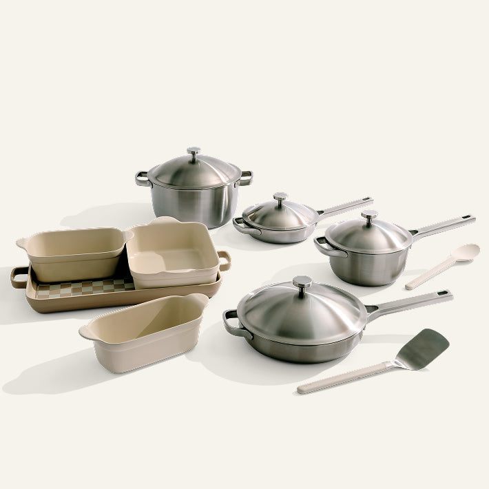 The Titanium Pro Cookware + Bakeware Set, featuring pots, pans, lids, beige ceramic baking dishes, a spatula, spoon, and nonstick bakeware, is neatly arranged on a light-colored surface.