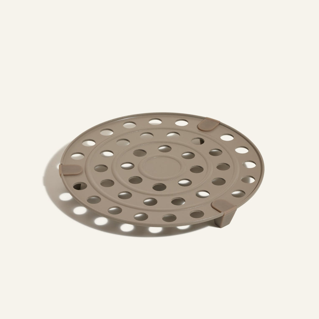The Perfect Pot Roasting Rack is a round beige plastic disc with multiple circular holes and three raised tabs, shown on a plain light background.