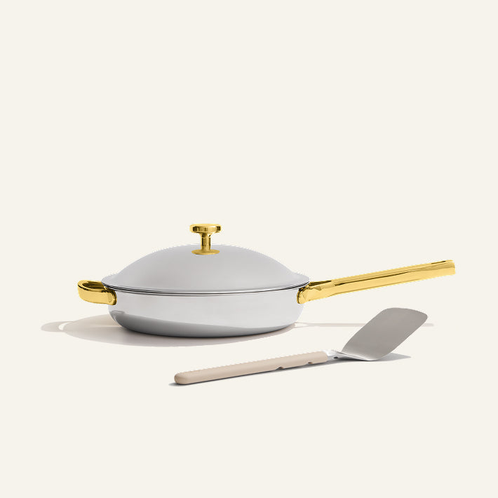 The Titanium Always Pan Pro, a white no-coating nonstick frying pan with a gold handle, sits on a light surface with a metal spatula placed in front.