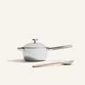 The Titanium Mini Perfect Pot Pro, a white non-toxic saucepan with lid and long handle, is placed beside a beige cooking spoon on a plain light background.
