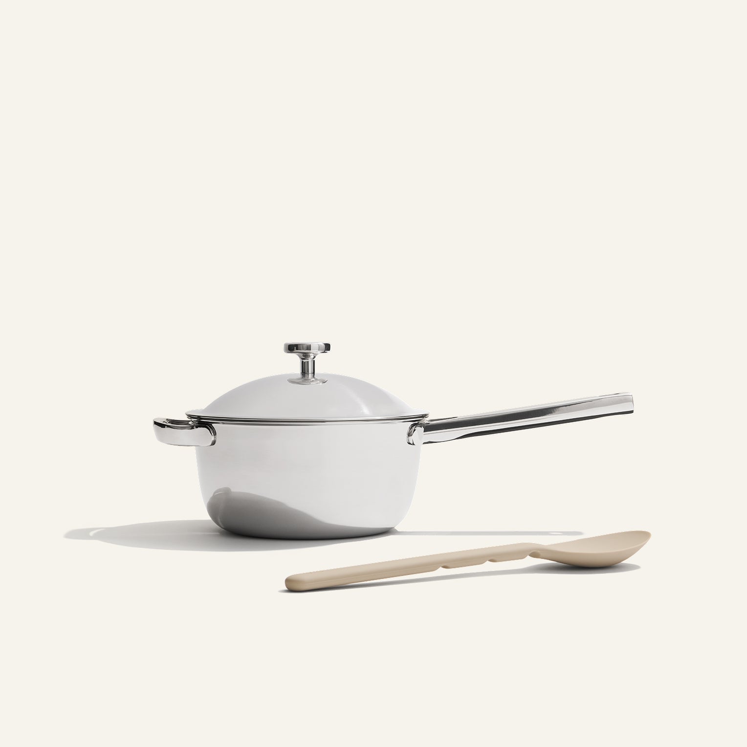 The Titanium Mini Perfect Pot Pro, a white non-toxic saucepan with lid and long handle, is placed beside a beige cooking spoon on a plain light background.