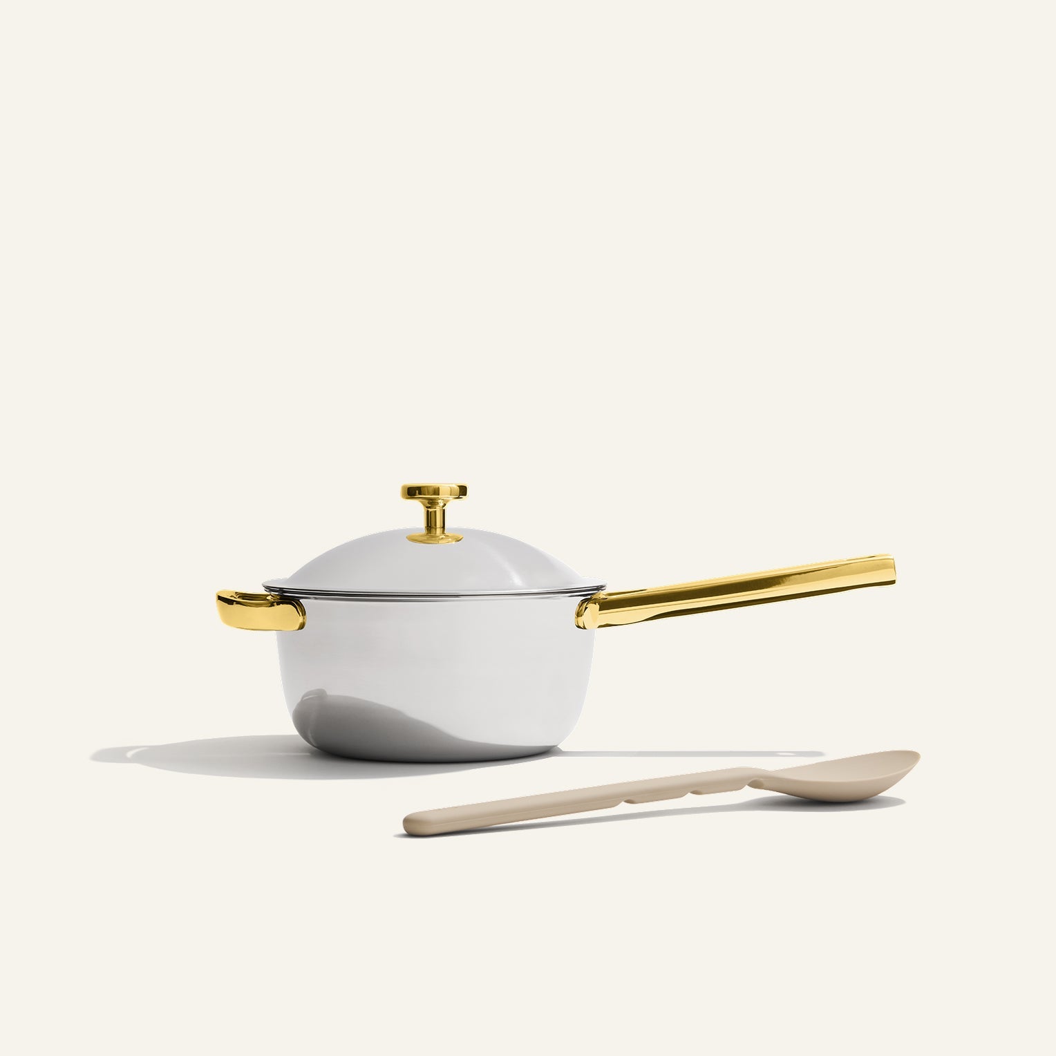 The Titanium Mini Perfect Pot Pro is a high-performance, non-toxic white saucepan with a gold handle and lid knob, paired with a beige spoon, displayed on a plain light background.