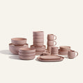 The Complete Stacking Set features matte blush pink ceramic bowls, plates, mugs, and oval platters stacked on a neutral backdrop—ideal as chic dinner party serveware or for stylish everyday hosting.